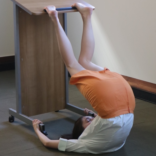 Photo of woman in orange skirt and white shirt with back on carpeted floor and body reaching up with feet onto a podium - experimental dance